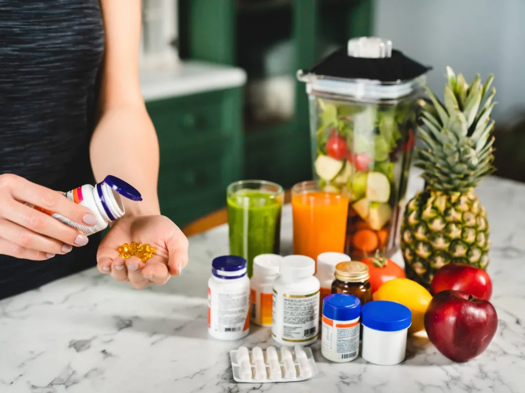 Supplements in different formats like capsules, tablets, fresh fruits, fruit juice and a smoothie near a blender filled with ingredients.